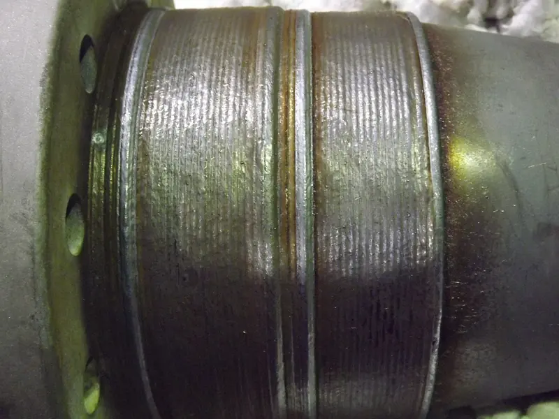 A close-up of ridged weld metal built up in bands around a steel cylinder, with a row of bolt holes at its edge.